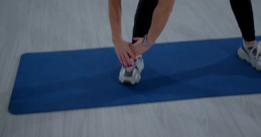 Bir kız, kapalı bir alanda mavi bir egzersiz minderinin üzerinde ayak bileğine uzanırken öne doğru eğilir. Esnekliği artırmak için esneme hareketlerine odaklanmış durumda..