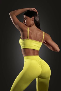 Genç bir kız sporcu güçlü bir duruşu olan sarı spor kıyafetlerle poz veriyor. Sahne karanlık bir geçmişi olan bir stüdyo. Atletik yapısını ve özgüvenini gösteriyor..