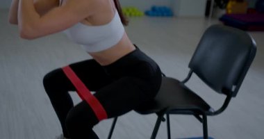 Genç bir kadın iskemlede otururken direniş bandını kullanarak oturarak çömelme hareketi yapıyor. Formuna odaklanıyor ve kaslarını bir spor salonunda çalıştırıyor..