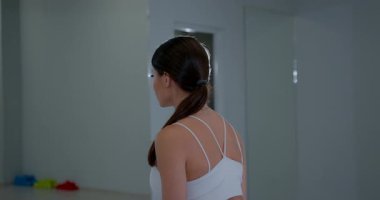 Spor salonunda duran bir kişi, dambıllarla sırayla dans ediyor. Etkinlik sırt kuvvetine odaklanıyor. Çevrede spor aletleri ve aynalar var. Gündüz vakti gibi görünüyor..