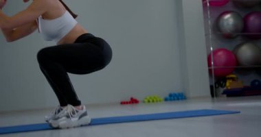 Kız spor salonundaki mavi bir paspasın üzerinde çömeliyor. Arka planda fitness ekipmanları yerleştirilirken o da egzersiz rutinine odaklanıyor. Işık boşluğu doldurur.
