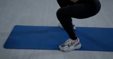 Kız içeride mavi bir egzersiz paspası kullanarak çömeliyor. Gücünü artırmak için egzersiz rutinine odaklanmış durumda. Bu sahne fitness ortamını vurguluyor.