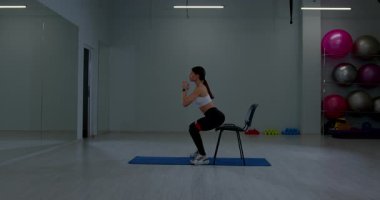 Genç bir kadın direniş bandı ve bir spor salonunda dambılla oturarak çömelme hareketi yapıyor. Döşeme temiz ve arka planda fitness ekipmanları görünüyor..