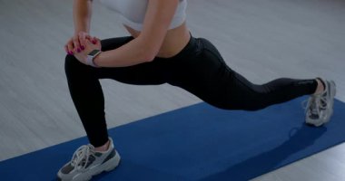 Genç bir kadın spor salonundaki mavi yoga minderine statik hamle yapıyor. Ön bacağı bükük ve arka bacağı düz iken ellerini birarada tutar..