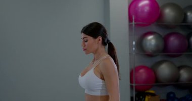Spor salonunda duran genç bir kadın statik hareketler yaparken vücuduna odaklanıyor. Spor kıyafetleri giyiyor ve fiziksel kondisyon ile meşgul..