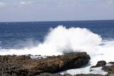Büyük bir dalga engebeli kıyıya çarpar. Arka planda okyanusun maviliği vardır..