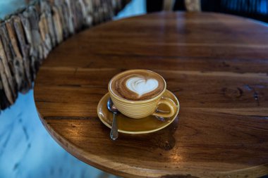Ahşap masa arkasında moda sarı kapuçino bardağı. Aşkın sembolü olarak kalp şeklinde latte sanatı. Sabah rutini için bir fincan..