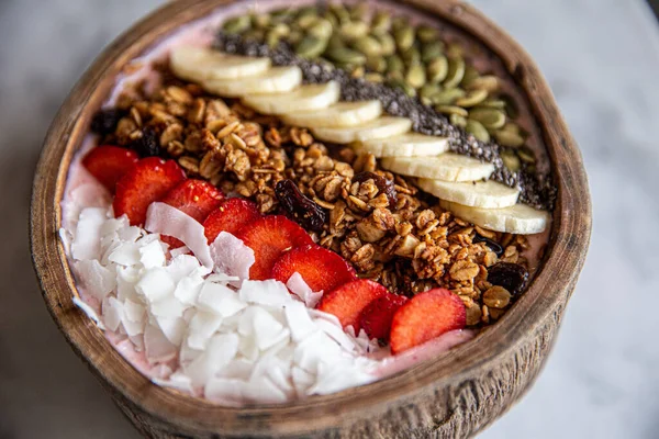 Kahvaltılık smoothie kasesi granola, çilek, hindistan cevizi, fındık, ayçiçeği, muz ve chia çekirdeği. Kase mermer masa arkasında.