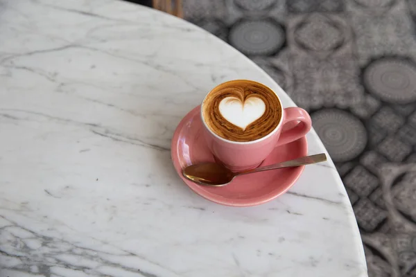 在大理石桌子的背景上 有时髦的粉色热卡布奇诺杯 心形拿铁艺术作为爱情的象征 每天早上喝一杯 图库图片