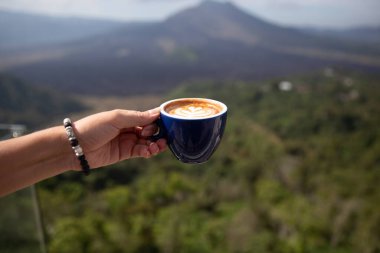 Dağ manzaralı bir kadının elinde bir fincan kapuçino, sabah vakti.