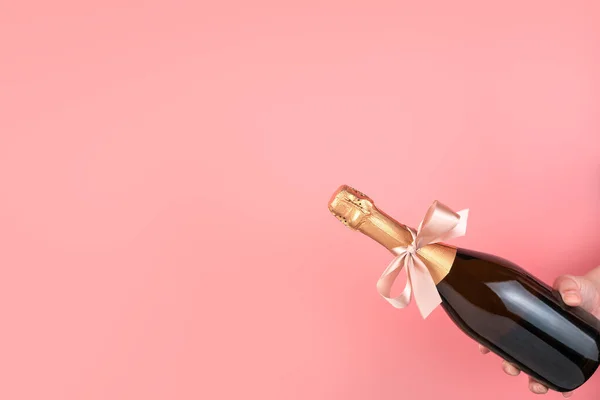 Uma garrafa de champanhe com um arco na mão sobre um fundo rosa. — Fotografia de Stock