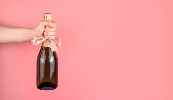 Mão segurando uma garrafa de álcool, decorada com um arco em um fundo rosa. — Fotografia de Stock