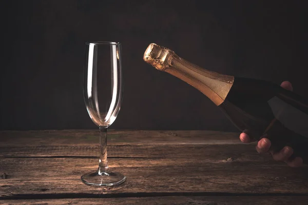 Uma mão segura uma garrafa de champanhe fechada ao lado de um copo de vinho vazio em um fundo de madeira. — Fotografia de Stock