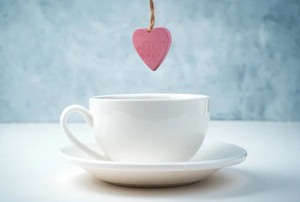 A pink heart hangs over a coffee Cup and saucer on a white and gray background. — Stock Photo, Image