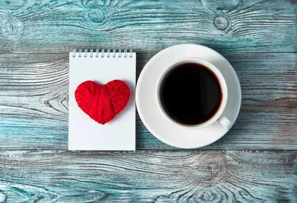 A red heart on a white Notepad and a Cup of coffee on a wooden gray-blue background. — Stock Photo, Image