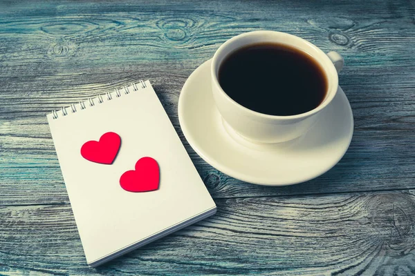 White coffee mug and two red hearts on a Notepad on a woodTwo red hearts on a white Notepad next to a coffee Cup on a wooden background.en background. — Stock Photo, Image