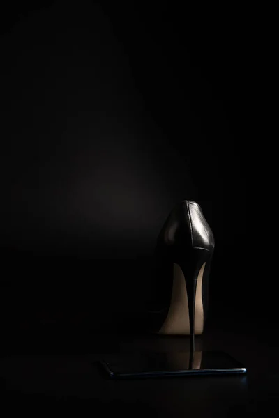 One womans high-heeled shoe and a phone on a black background.