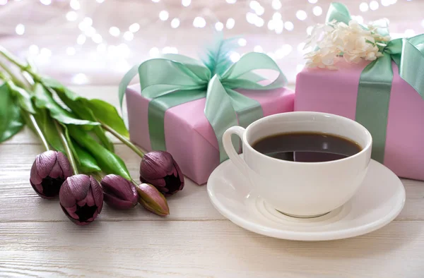 Fondo atmosférico festivo con una taza de café, flores y envases de regalo en el fondo de una guirnalda en llamas. —  Fotos de Stock