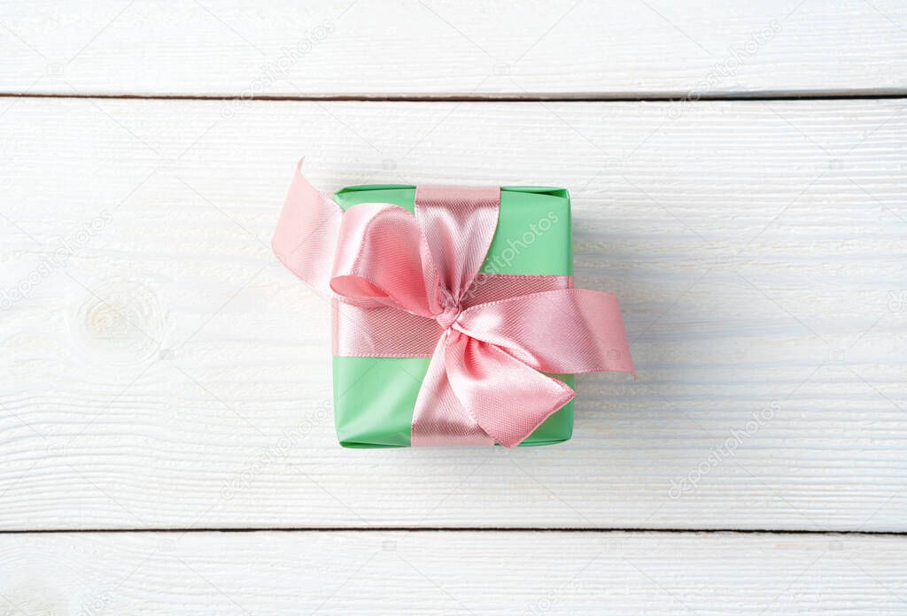 Green gift box with pink ribbon on white wooden background.