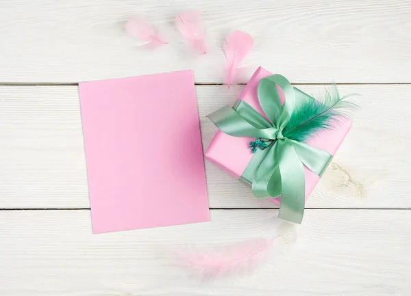 Um envelope rosa e uma caixa de presente com penas em um fundo de madeira leve. — Fotografia de Stock