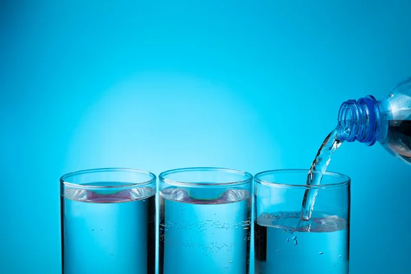 Woda w szklankach, napełnianie trzech szklanek z plastikowej butelki na niebieskim tle. — Zdjęcie stockowe