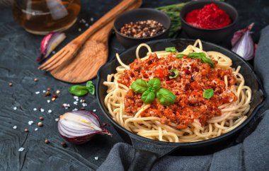 Kızartma tavasındaki spagetti ve sos üzerine peynir ve taze fesleğen serpilmiş siyah beton arka planda baharat ve soğan arkaplanı..