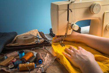 Kadın eli olan parlak bir dikiş, sarı kumaş, iplik ve dikiş makinesi mavi arka planda. Kopyalama alanı olan yan görünüm.