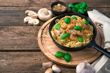 Frying pan with pasta with mushroom sauce on a cutting board with a background of mushrooms and garlic. Side view, horizontal. clipart