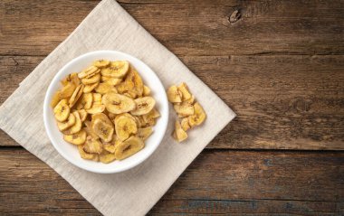 Ahşap bir arka planda, beyaz bir tabakta kurumuş muz parçaları. Üst görünüm, yakın çekim.