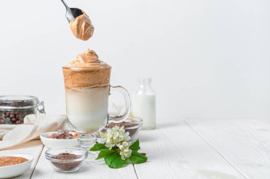 Dalgona 'nın buzlu kahvesi ve köpük kaşığı beyaz arka planda, içerik ve renklerle birlikte. Yan görünüm, yatay.