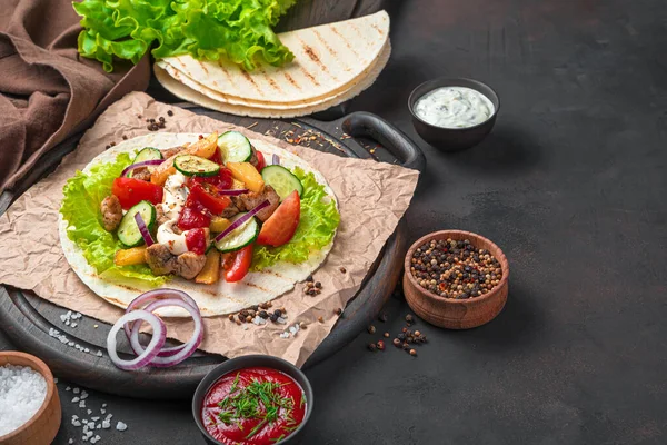 Meat, fried potatoes, vegetables, salad and sauce on a thin pitta on a brown cutting board. Preparation of tacos, shawarma, burritos. Side view, copy space.