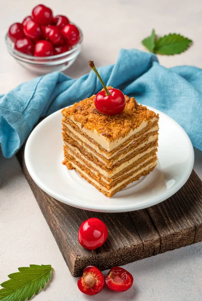 A piece of honey cake decorated with fresh cherries on a light background. Side view, vertical.