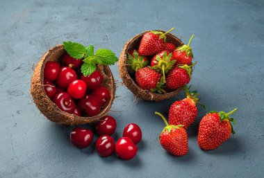 İki hindistan cevizi kabı sulu, olgun kirazlar ve koyu mavi arka planda çileklerle doludur. Yan görünüm, yakın plan.