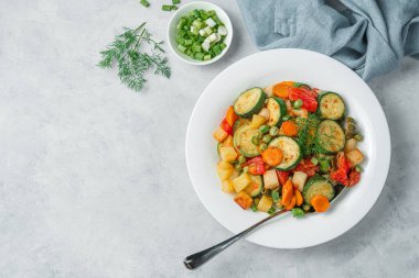 Sağlıklı sebze güveci, gri arka planda beyaz bir tabakta, yeşilliklerle birlikte. Üst görünüm, alanı kopyala.