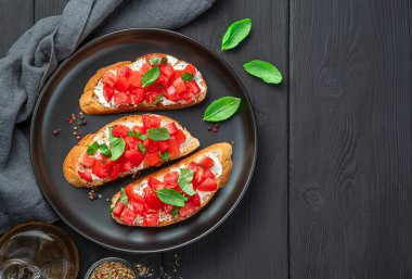 Siyah ahşap arka planda domates, fesleğen, tereyağı ve peynirli İtalyan bruschetta 'sı. Üst görünüm, boşluğu kopyala. Domatesli kızarmış ekmek..