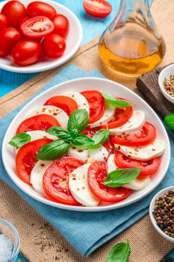 Domatesli Caprese salatası, mozzarella ve mavi arka planda fesleğen. Yan görünüm, dikey.