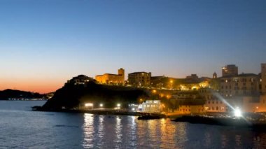 Şehir ışıkları ve deniz feneriyle birlikte Piombino 'nun promontuarının sabit gerçek zamanlı görüntüleri günbatımında, İtalya' da denizin sakin sularını yansıtıyor.