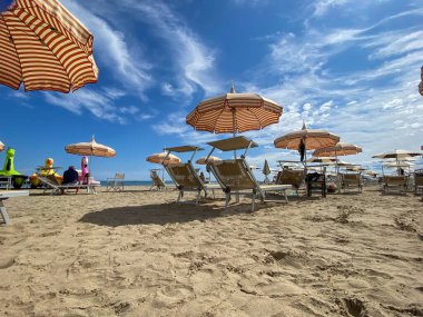 Low-angle, bright sun-drenched view of a sandy beach in Cesenatico, featuring rows of striped umbrellas and empty deck chairs under a dramatic blue sky with wispy clouds. High quality photo