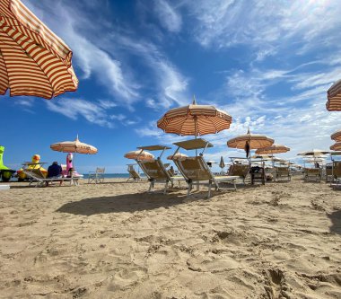 Low-angle, bright sun-drenched view of a sandy beach in Cesenatico, featuring rows of striped umbrellas and empty deck chairs under a dramatic blue sky with wispy clouds. High quality photo