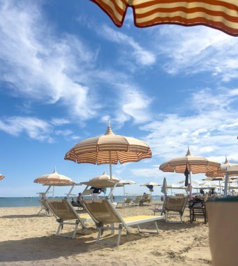 Low-angle, bright sun-drenched view of a sandy beach in Cesenatico, featuring rows of striped umbrellas and empty deck chairs under a dramatic blue sky with wispy clouds. High quality photo