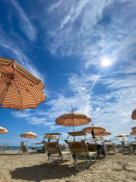 Low-angle, bright sun-drenched view of a sandy beach in Cesenatico, featuring rows of striped umbrellas and empty deck chairs under a dramatic blue sky with wispy clouds. High quality photo