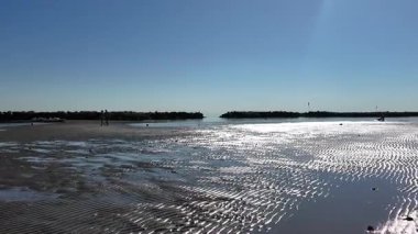 Wide view of Cesenatico beach at low tide, showing wet sand patterns and shallow water. People walk along the shore near modern buildings and a long breakwater under clear blue sky. High quality 4k footage