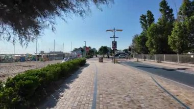 A dynamic ground-level view of a paved seaside promenade in Cesenatico with modern architecture, lush green shrubs, and contemporary street lights under a clear blue sky. High quality 4k footage
