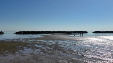 Wide view of Cesenatico beach at low tide, showing wet sand patterns and shallow water. People walk along the shore near modern buildings and a long breakwater under clear blue sky. High quality 4k footage