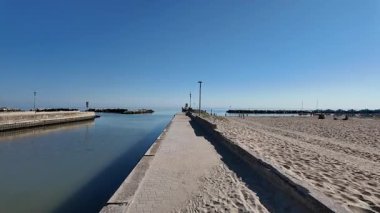 A concrete jetty separating a sandy beach from the calm, blue water of a canal entrance or harbor mouth in Cesenatico. The scene is dominated by a clear blue sky on a sunny day. High quality 4k footage