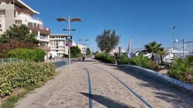 A dynamic ground-level view of a paved seaside promenade in Cesenatico with modern architecture, lush green shrubs, and contemporary street lights under a clear blue sky. High quality 4k footage