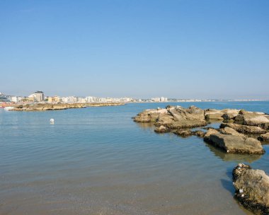 Sakin denizde, insan yapımı, engebeli bir kaya dalgasının alçak açılı görüntüsü. İtalya 'nın Cattolica tatil beldesinin modern ufuk çizgisi açık mavi gökyüzünün altında arka planda görünür.. 