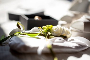 Soft-focus, bright image of a white satin ring bearer pillow with a delicate white rose and green ribbon, next to an open, black jewelry box. High quality photo