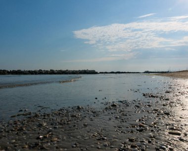 A ground-level view of a wet, pebbly beach foreground leading to a calm sea and a long line of rock breakwaters under a blue sky with white clouds. High quality photo