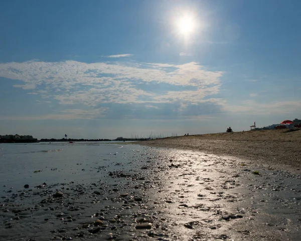 A ground-level view of a wet, pebbly beach foreground leading to a calm sea and a long line of rock breakwaters under a blue sky with white clouds. High quality photo